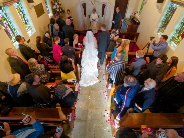 Bride walking down aisle at Saint Oswald's Memorial Church heritage wedding venue ceremony with guests seated in wooden pews and stained glass windows