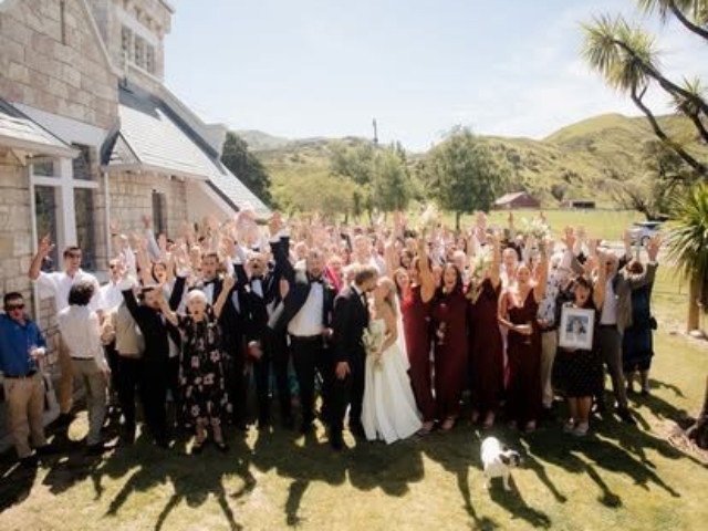 Large wedding party gathered on lawn outside historic stone church with mountains in background, celebrating heritage venue ceremony