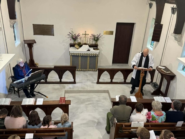Interior of Saint Oswald's Memorial Church with congregation seated in wooden pews during a heritage chapel ceremony, featuring historic architecture and stained glass windows.