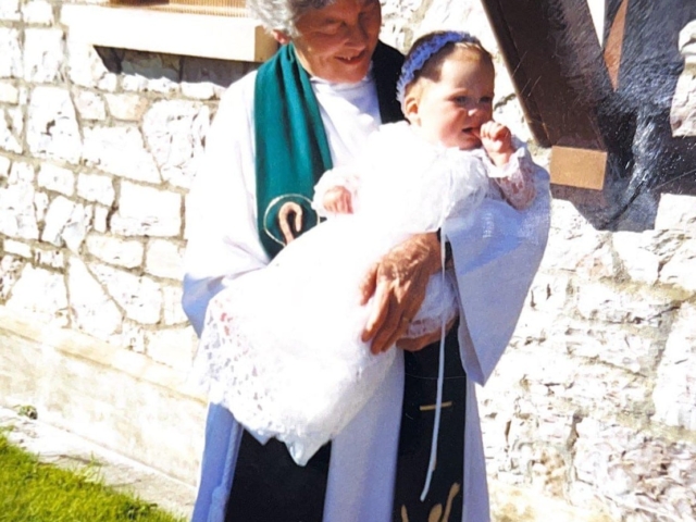 Woman holding infant in christening gown outside historic stone church, heritage chapel venue with traditional architecture and green grounds